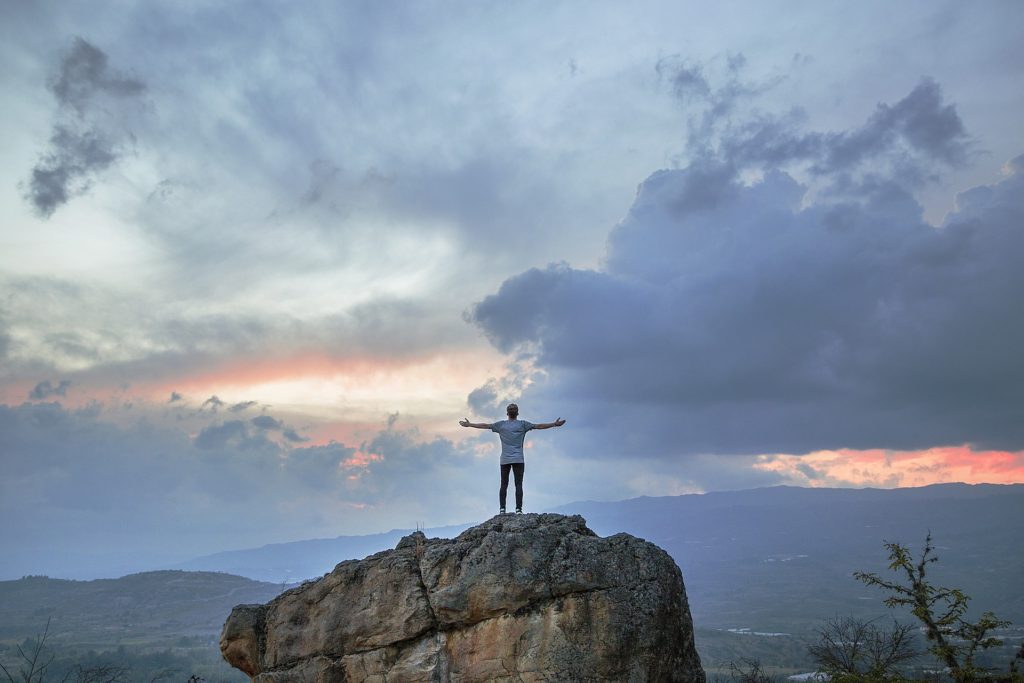 Photo of a person with outstretched arms, standing on a huge rock facing sunset