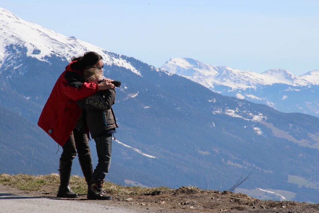 Mom with arms around her child helping her view the mountains through binoculars.