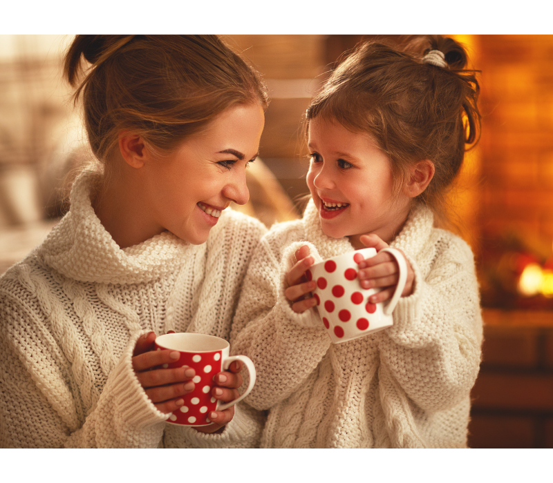 Mom and little girl, in matching sweaters, are holding mugs and smiling.