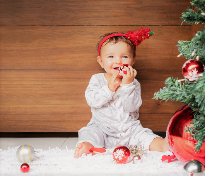 Baby in pajamas holding red ornament beside Christmas tree.