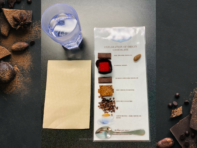Shows the laminated card with various forms of chocolate on it, a spoon, a napkin, and a glass of water. Photo taken by Sally Matheny at the French Broad Chocolate Factory, 2022.